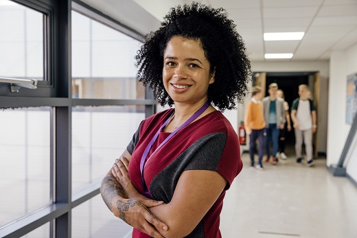 Portrait of a mixed race teacher with tattoos in the school hall. She has her arms folded and is smiling for the camera.