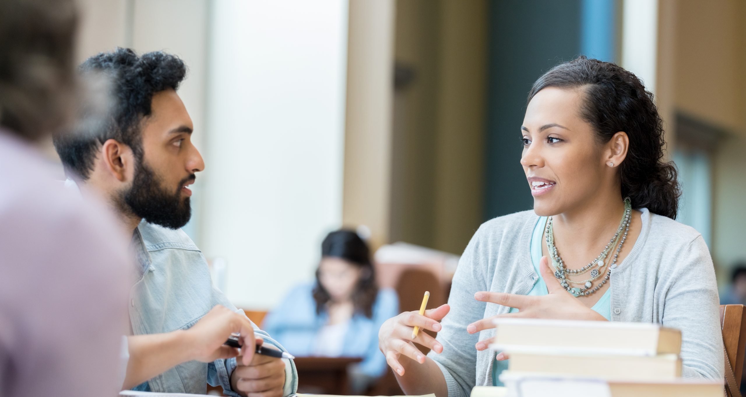 Two adults talk at a table with books and other people in the background