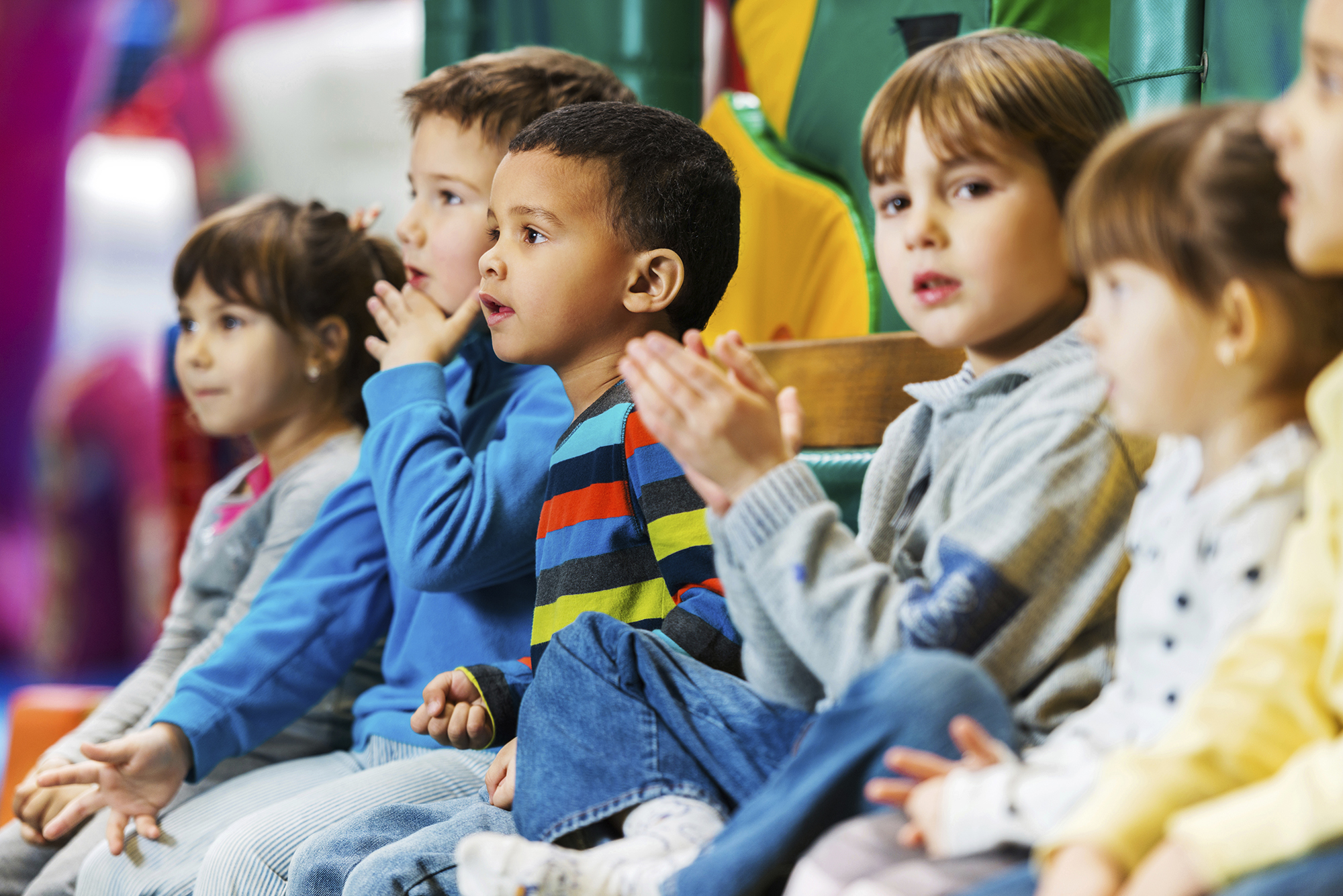 Group of young children sitting on the floor looking towards someone. One child is clapping.