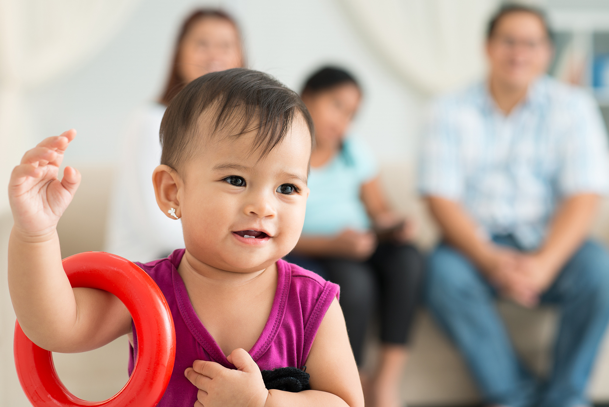 Toddler stands with a red ring toy around her arm. Her family is out of focus in the background. 