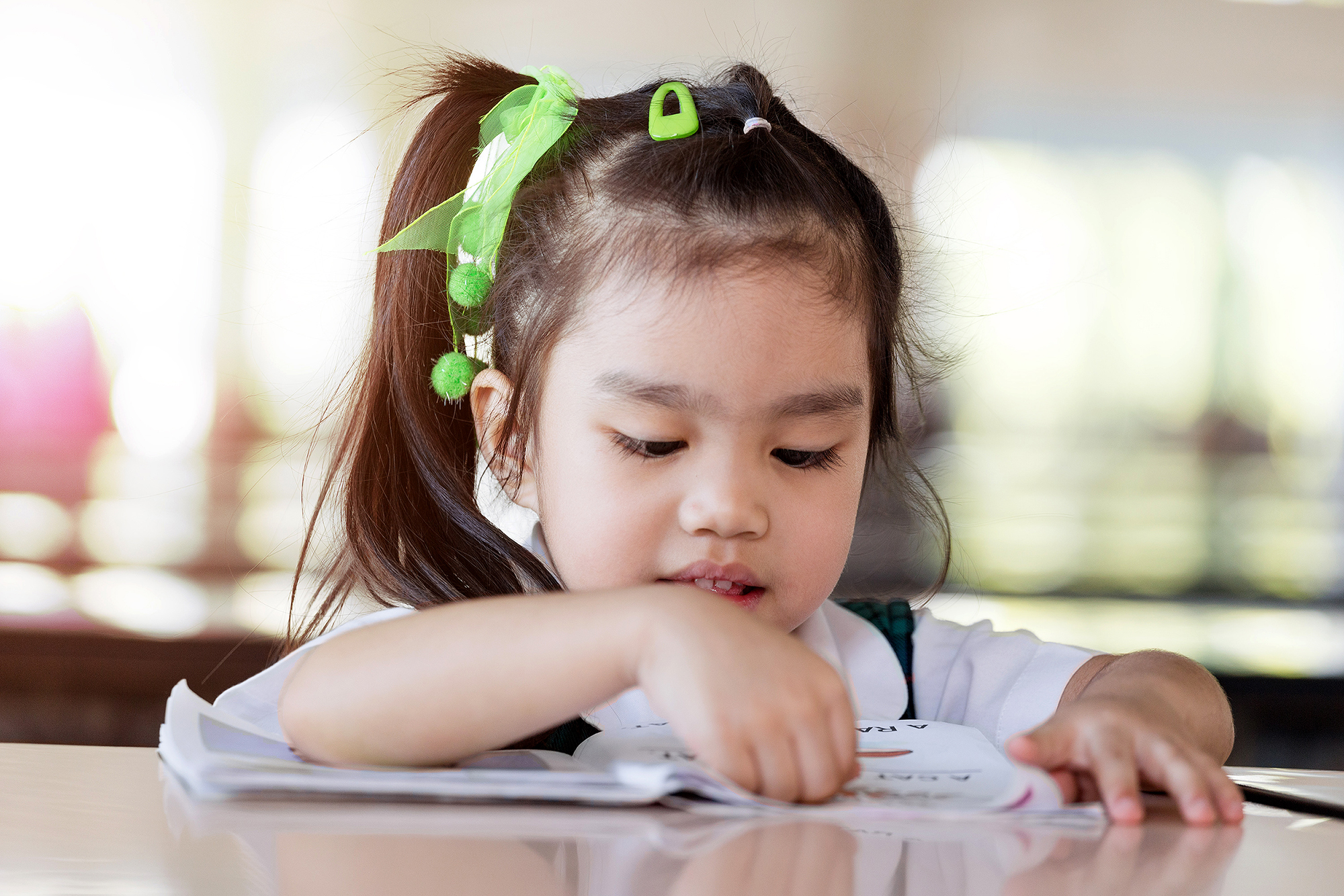 Young Asian child with green bows in her hair sits at a table reading.