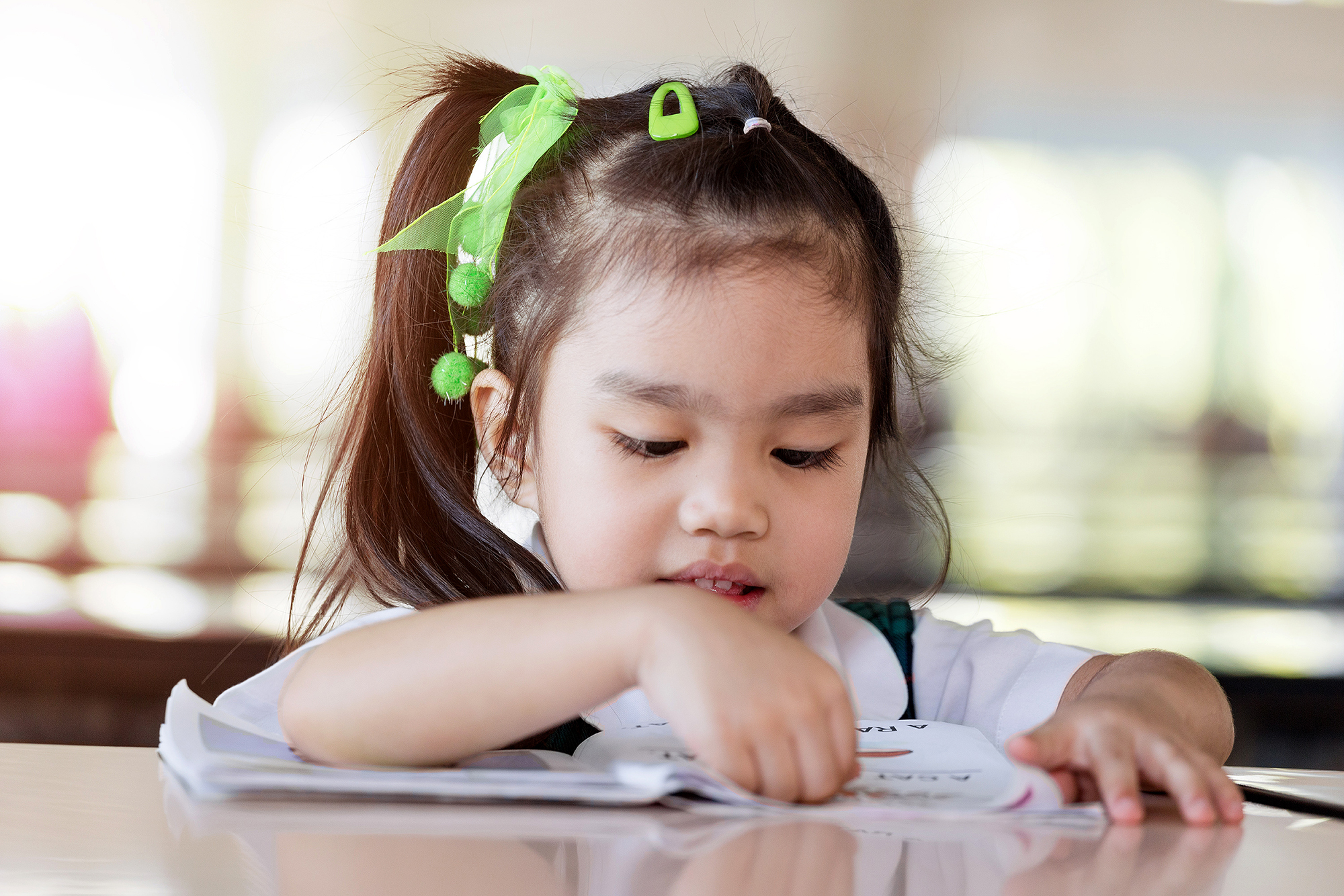 education and school concept asian (japan, chinese, korea) prett Young Asian child with green bows in her hair sits at a table reading.