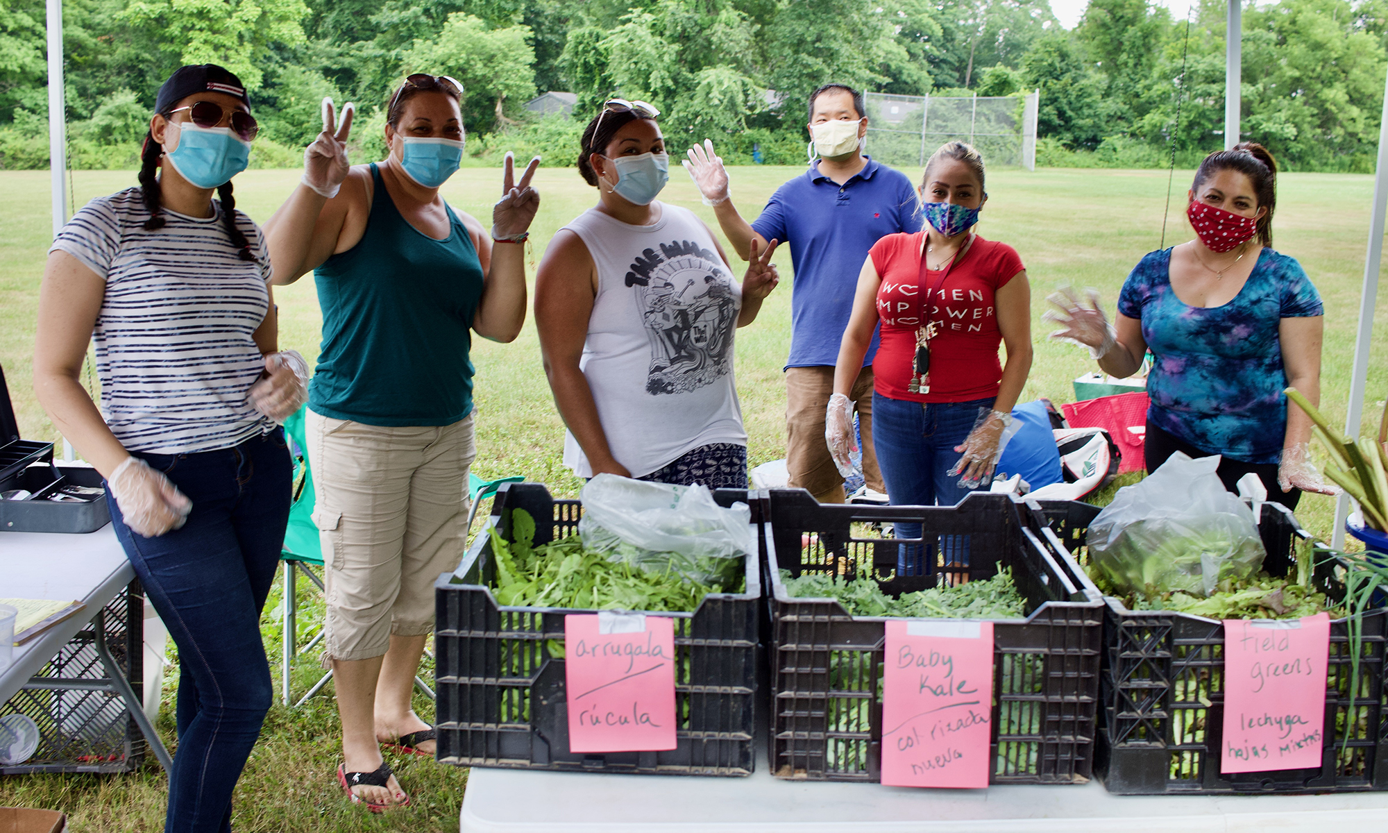 Group of adults standing in a line, outside. They are wearing masks. There are vegetables in bins on the table in front of them. 