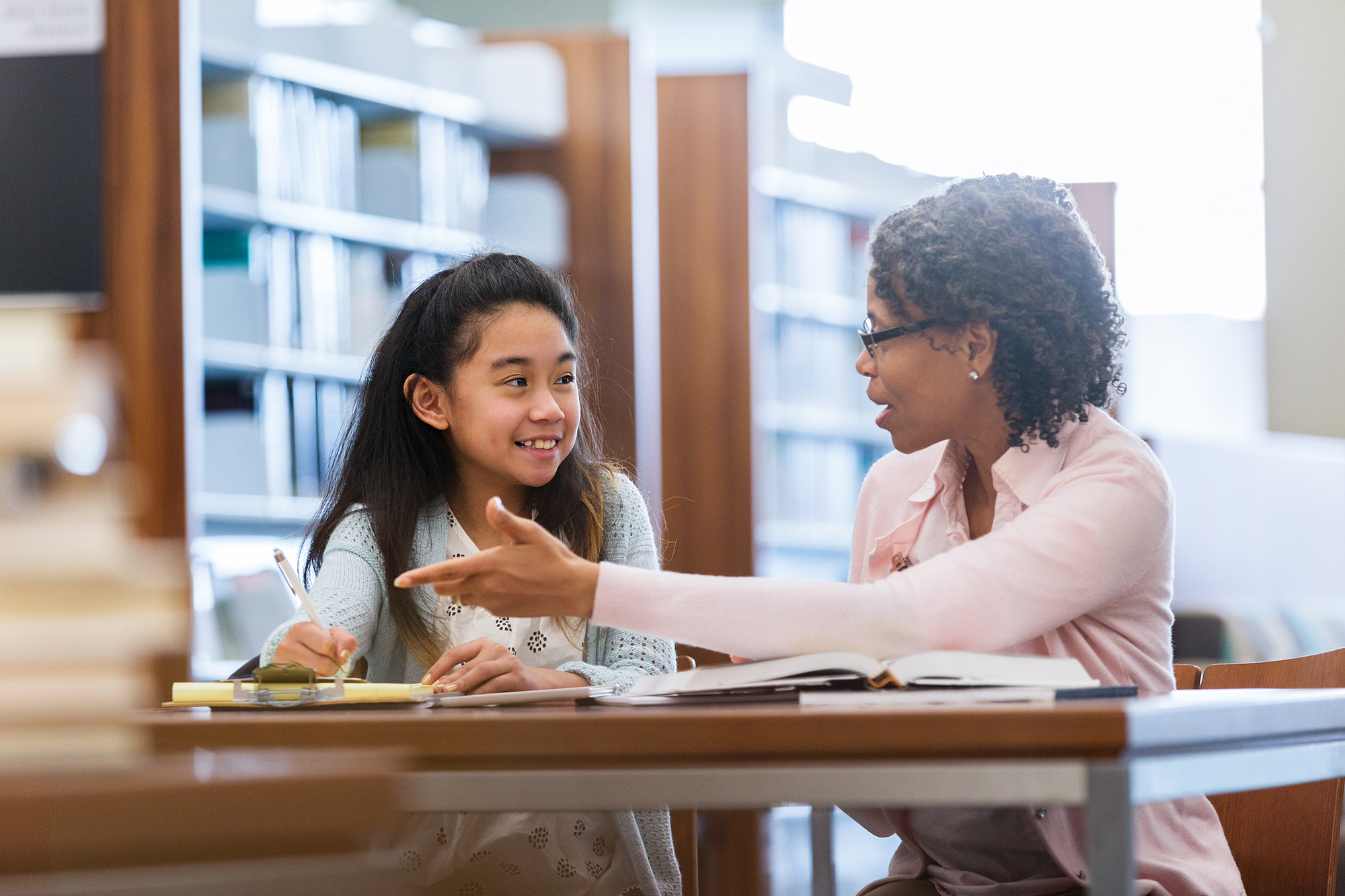 A smiling elementary age girl sits with her mature female mentor in a school library.  She enjoys a conversation.