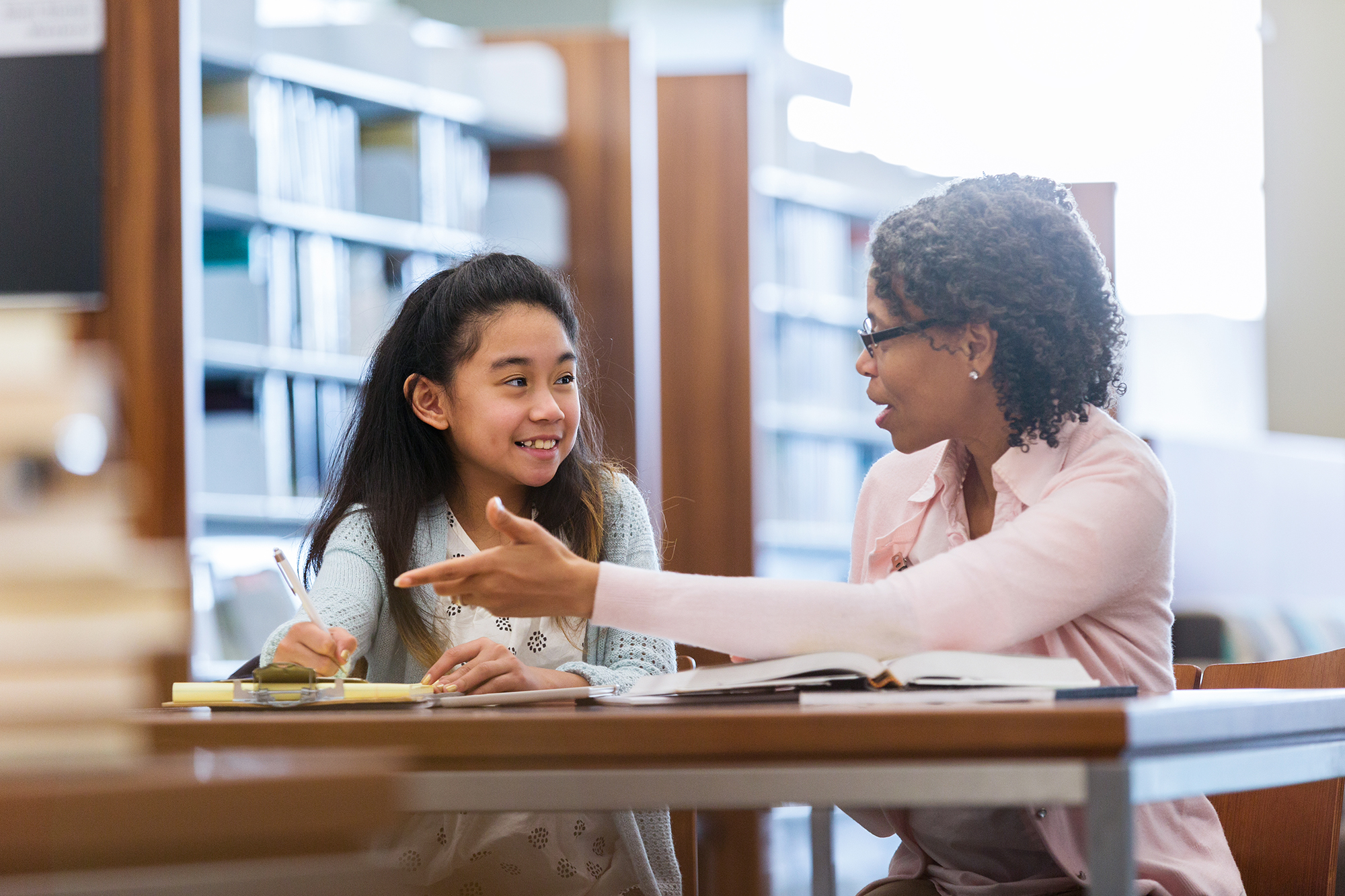 Elementary age girl receives help from mentor A smiling elementary age girl sits with her mature female mentor in a school library. She enjoys a conversation.