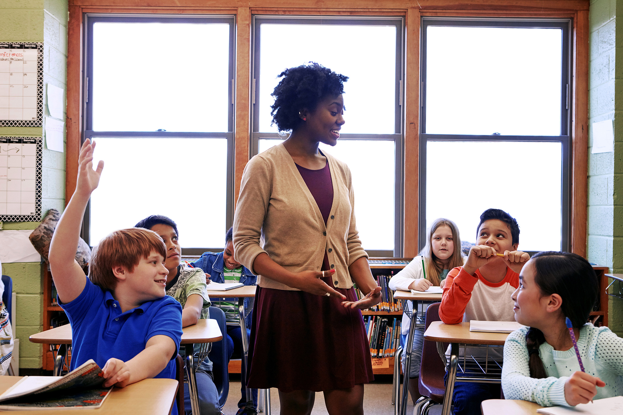 African American teacher talking to students who are sitting at desks. One student has his hand raised.