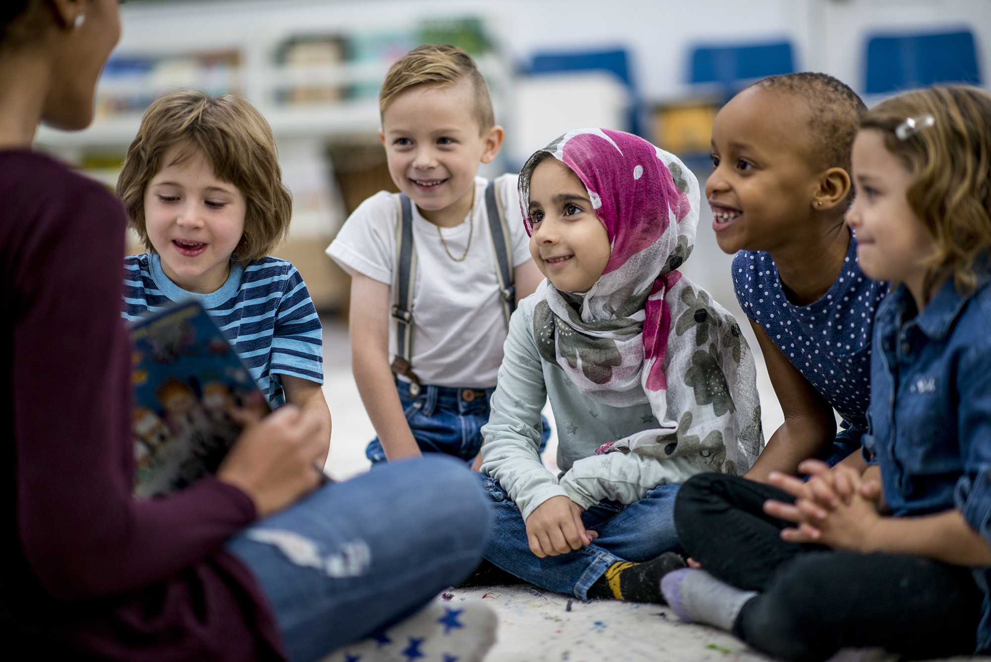 Reading A Storybook A multi-ethnic group of school children are indoors in a classroom. They are wearing casual clothing. They are sitting on the floor and eagerly listening to their teacher read a storybook.