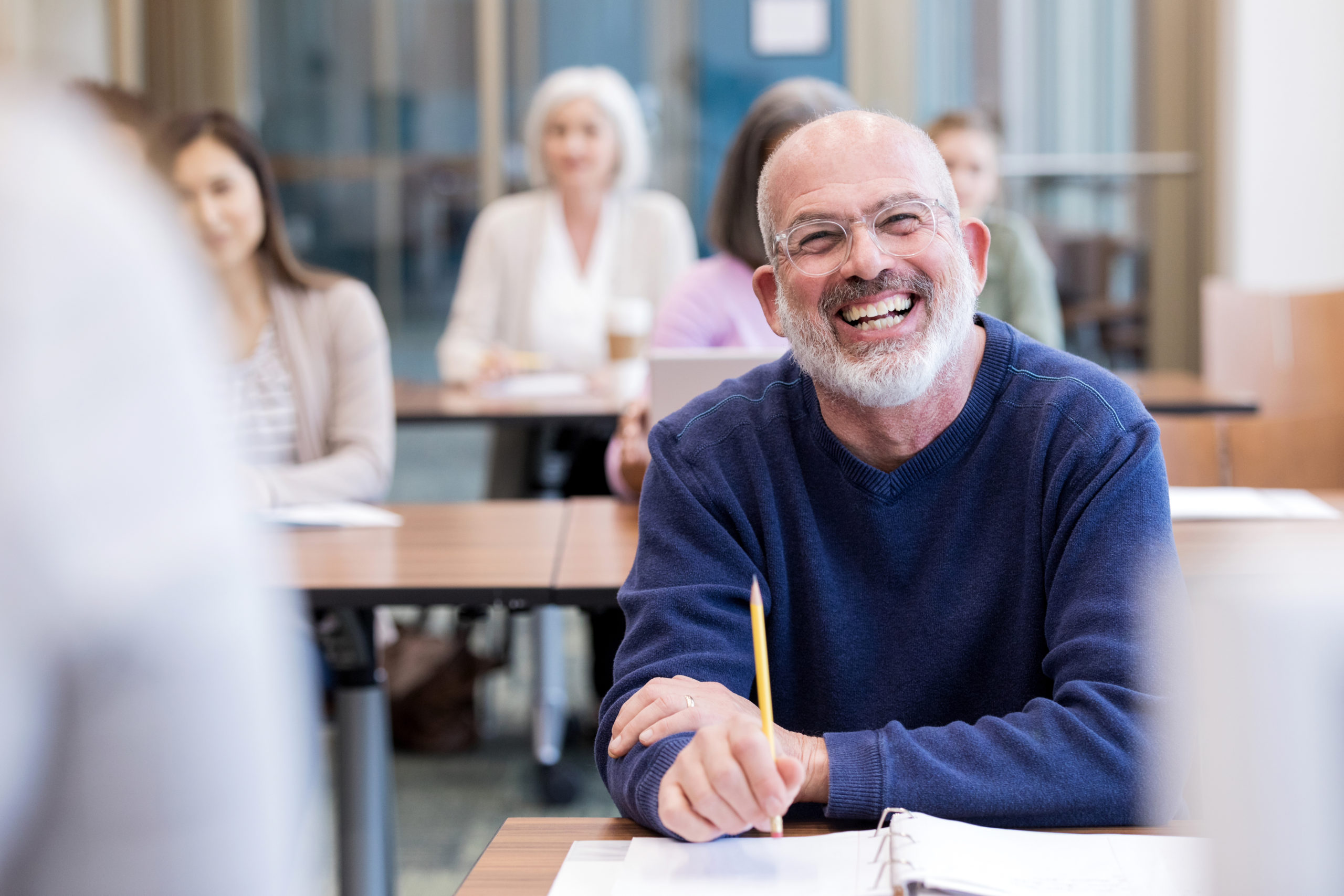 person with grey hair smiling and holding a pencil, in a room with other people taking a class