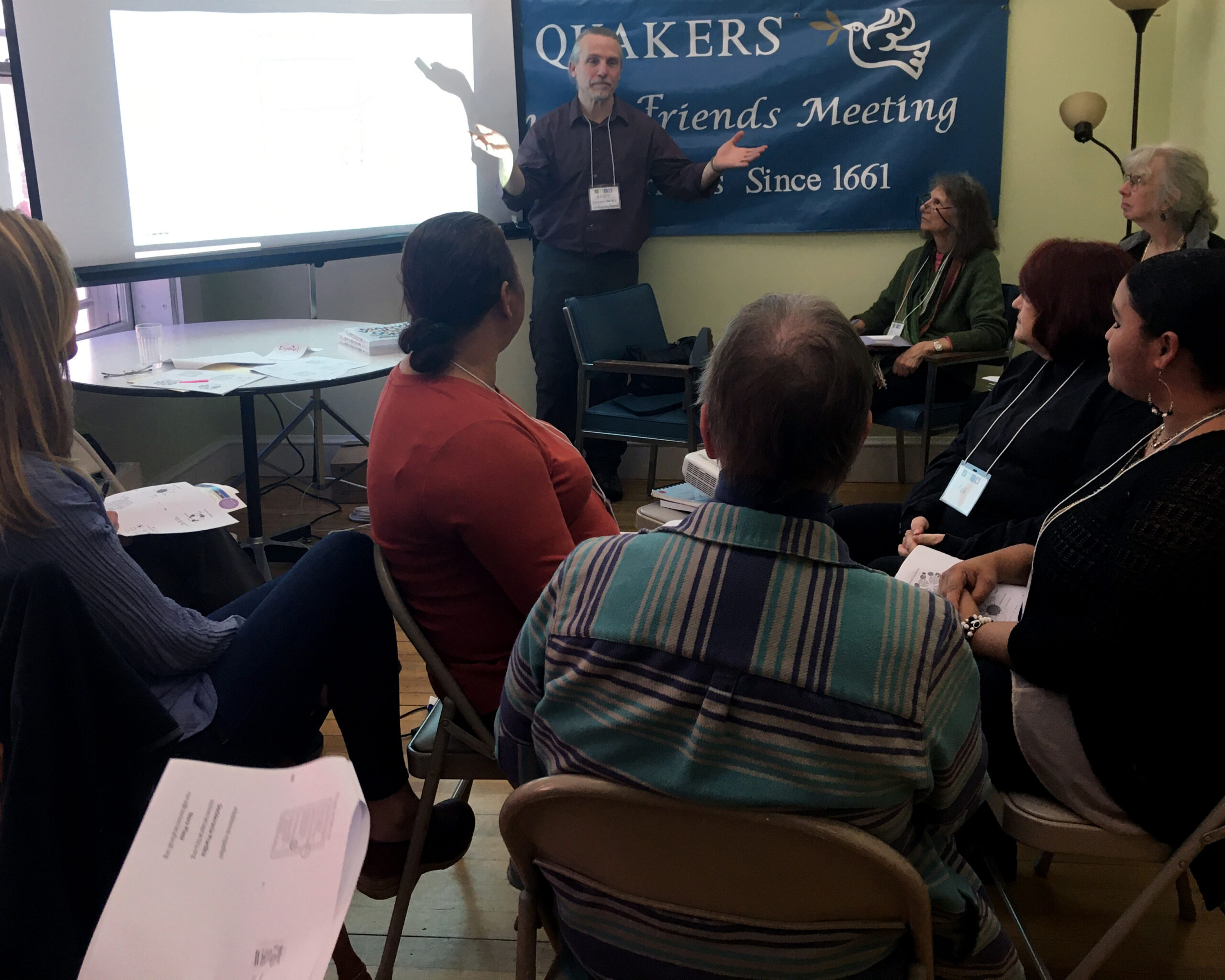 person presenting in front of a projector screen to a room full of seated people