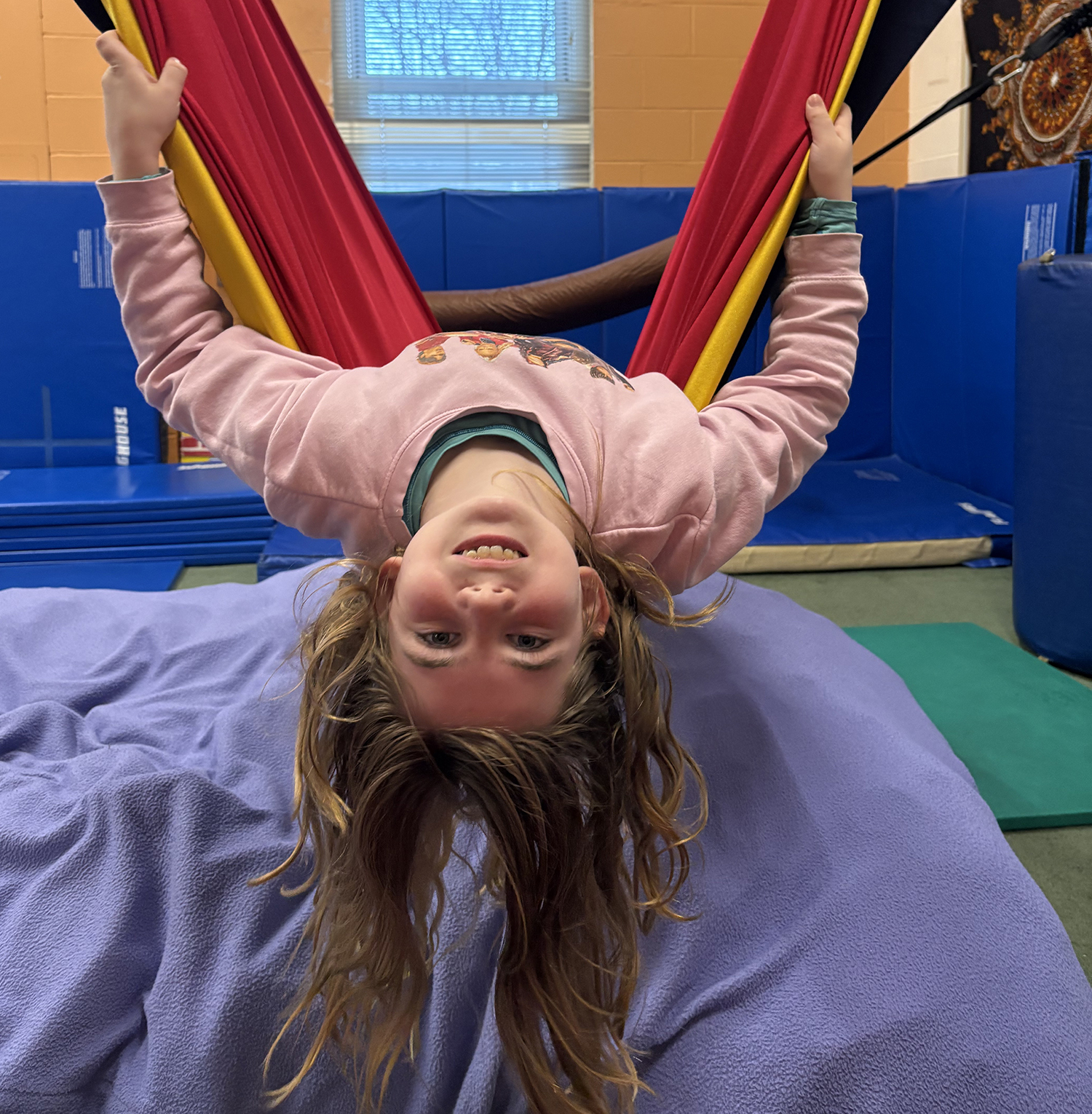 Little girl hanging upside down from a hamock, with her long blond hair hanging down. There are blue cushions underneath her. 