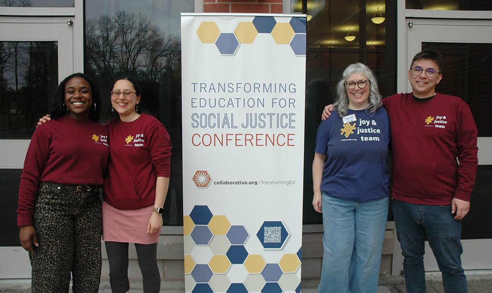 Four people standing in a line with their arms around each other. In the middle is a tall banner that reads Transforming Education for Social Justice Conference.