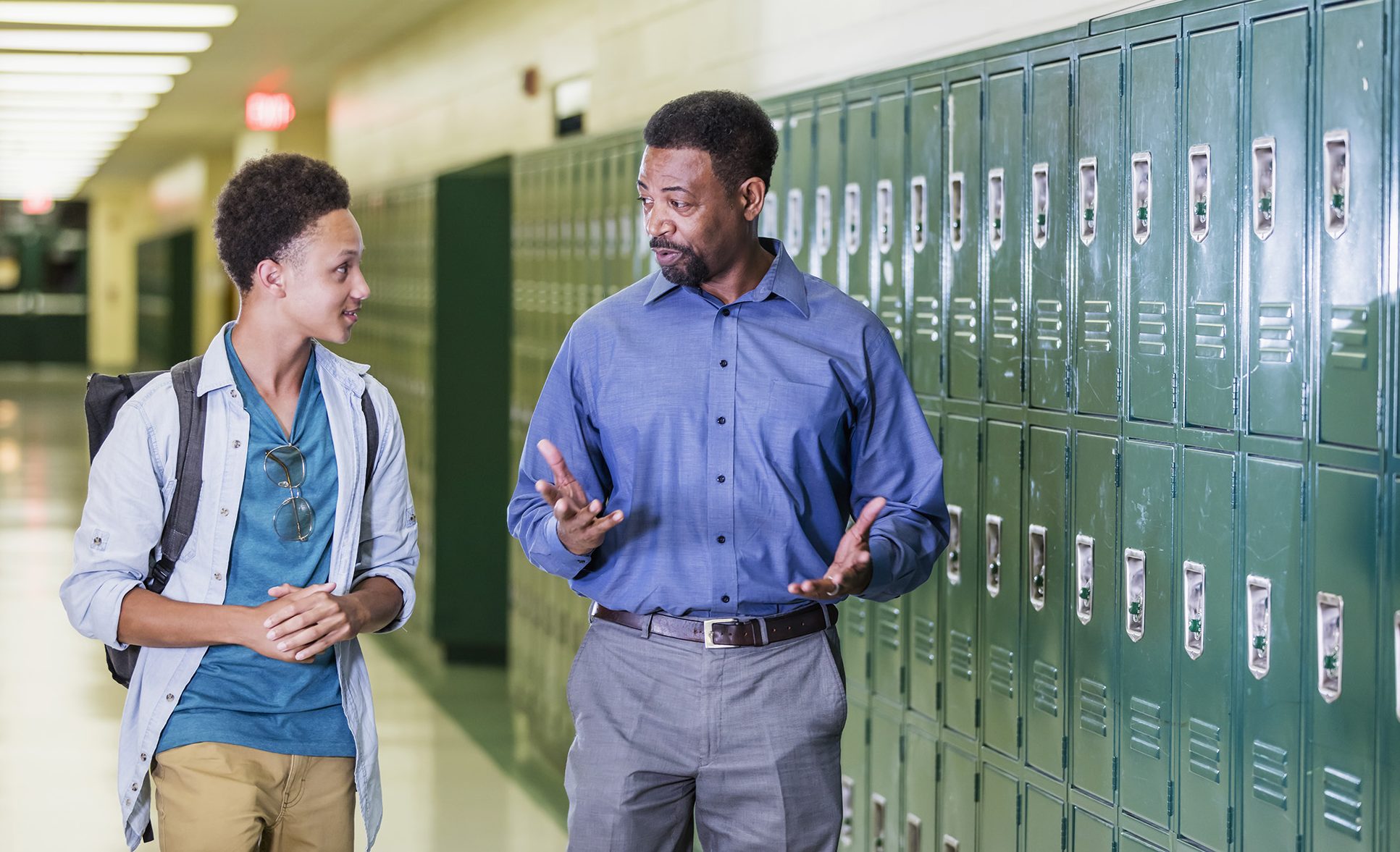 An African-American high school student walking with his teacher or school principal, in the hallway by a row of lockers. They are talking. The teacher has a serious expression on his face, a mature man in his 50s. The male student is an 18 year old teenage boy carrying a backpack.