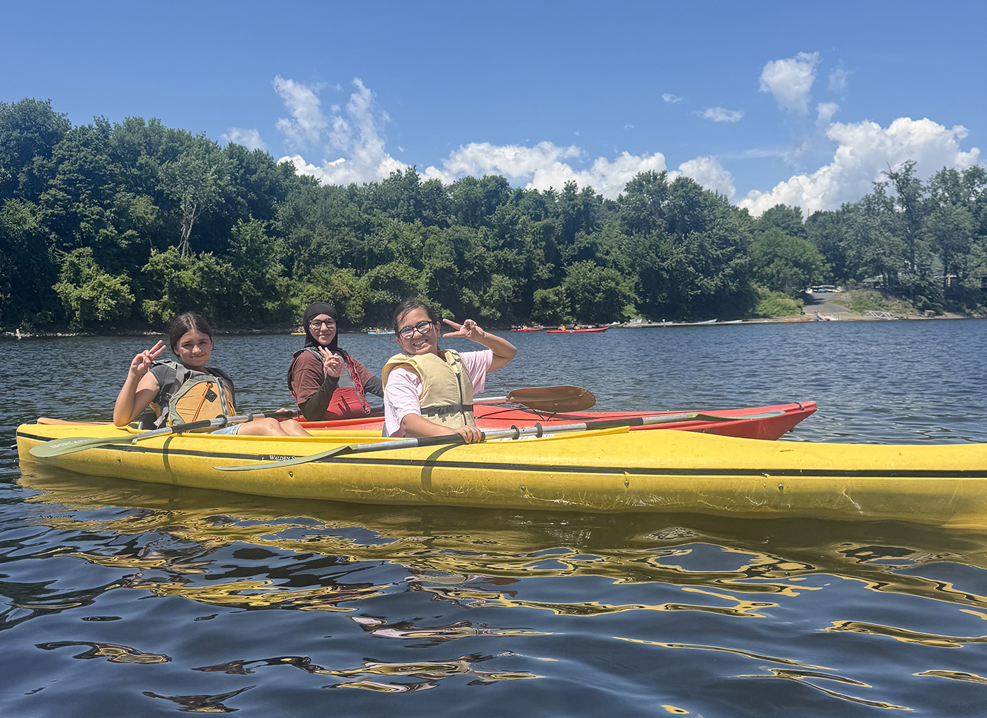 Three students in kayaks smiling at the camera and making peace signs. They are on the water with blue sky and trees in the background. 